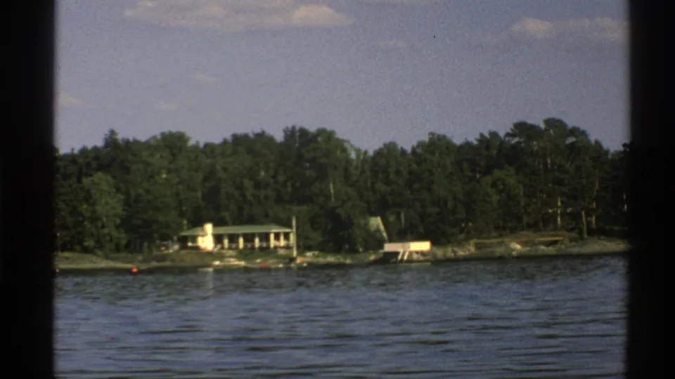 1976 Norway 8mm frame showing a pale house with green roof on a wooded rocky shoreline across the lake.