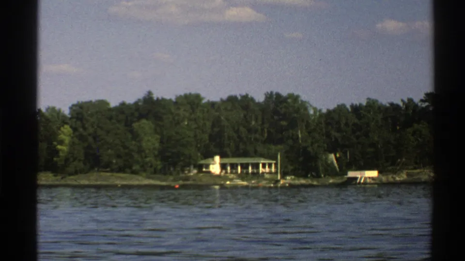 1976 Norway 8mm frame showing a lakeside home partly framed by forest, with dock features and a buoy near shore.