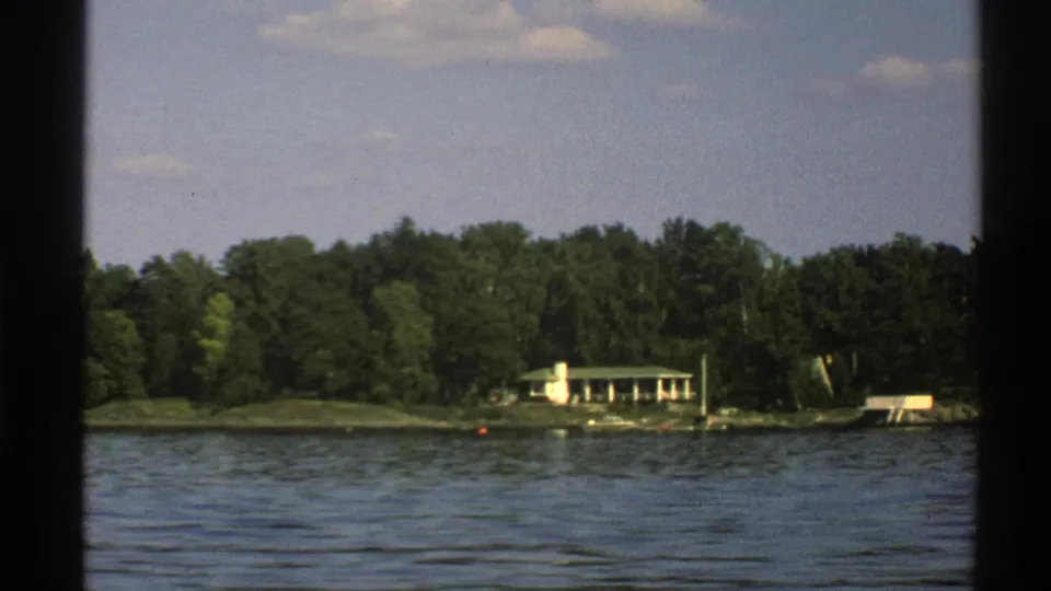 1976 Norway home-movie frame with the house shifted right in view, lake water in front and dense trees behind.