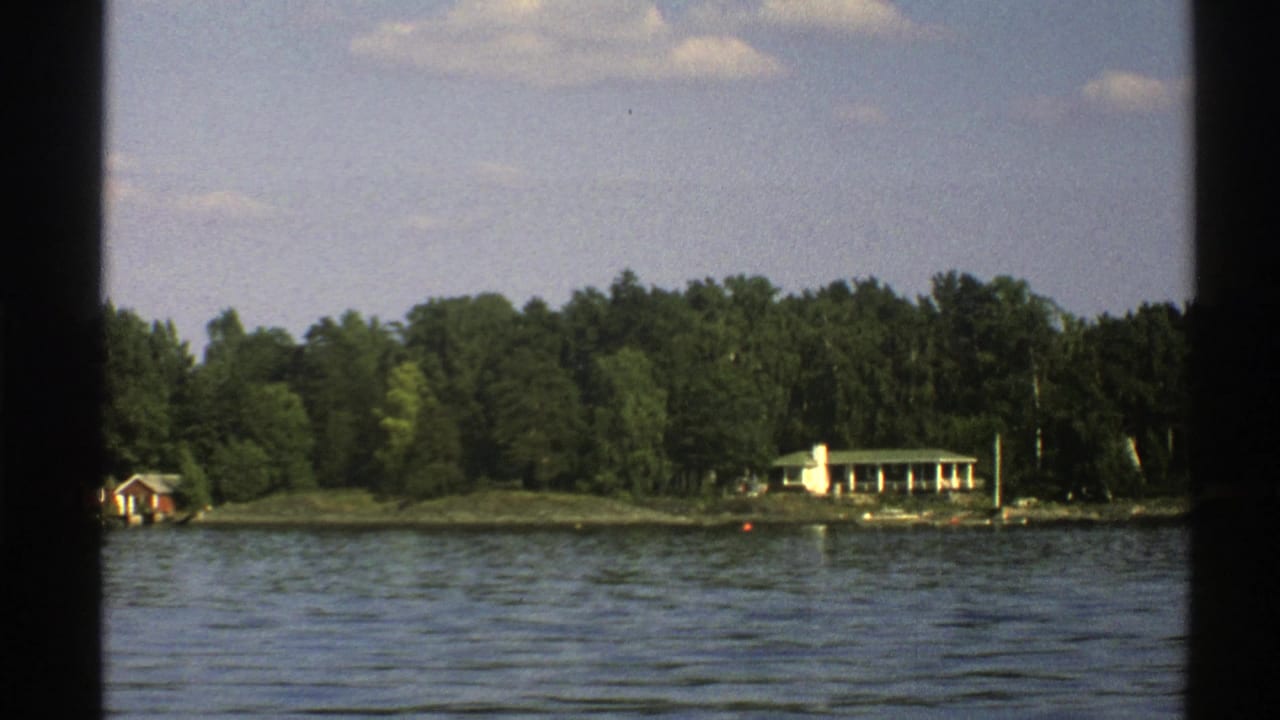 Forested shoreline with a white cottage, small red cabin, lake water, and dark film borders.