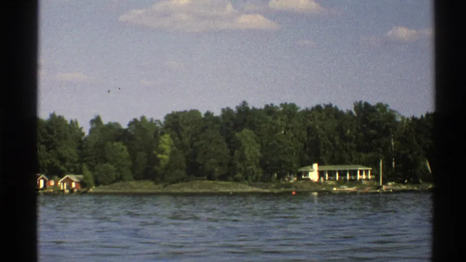 1976 Norway 8mm frame showing red shoreline cabins, a white cottage, dense trees, and blue lake water.