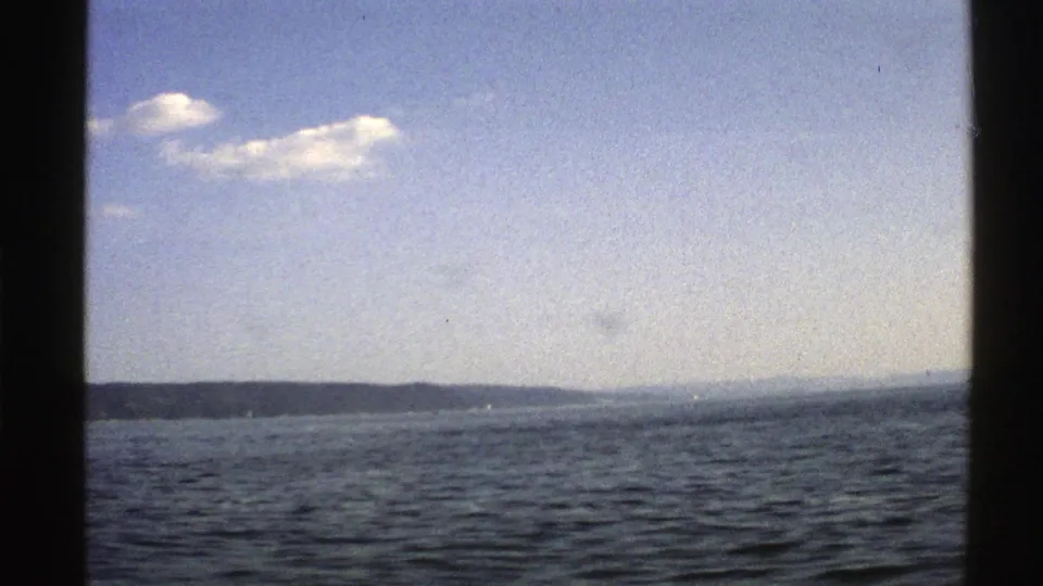 1976 Norway 8mm frame widening to open lake water, distant wooded horizon, and a partly cloudy sky.
