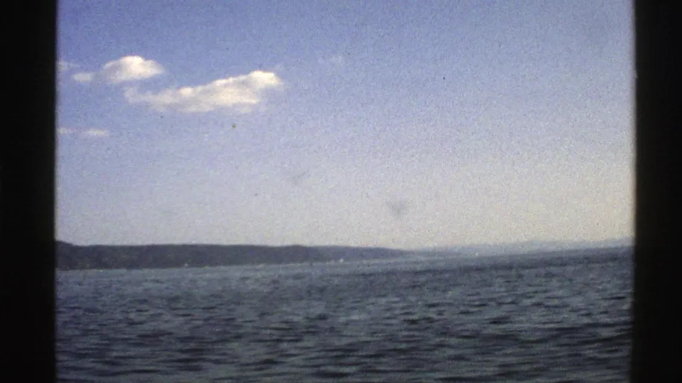 1976 Norway 8mm frame showing open water, a distant peninsula, pale blue sky, soft clouds, and dark frame edges.