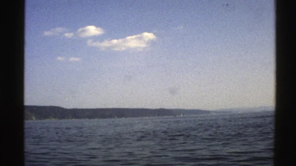 Norway 1976 home-movie frame of open sea, distant coastal land, bright sky, clustered clouds, and visible 8mm texture.