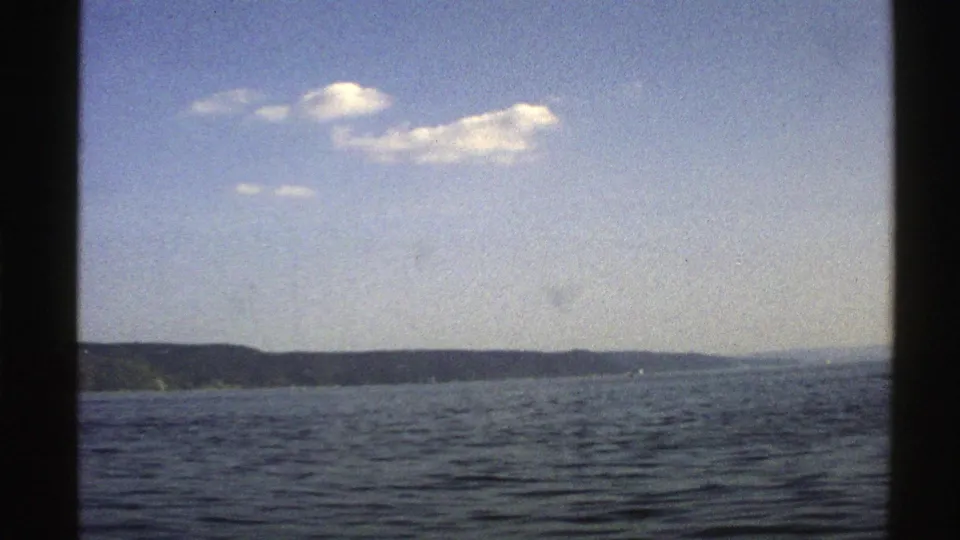 1976 Norway 8mm view of a broad water surface, dark low shoreline, pale sky, and soft clouds from a moving travel vantage point.