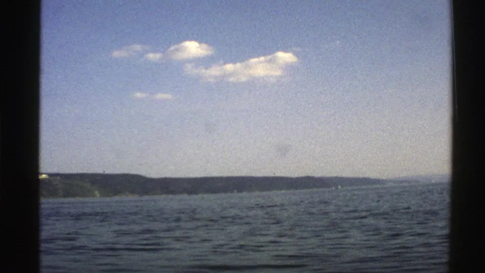 1976 Norway 8mm frame showing calm water, distant green shoreline, blue sky, and dark gate edges from a boat viewpoint.