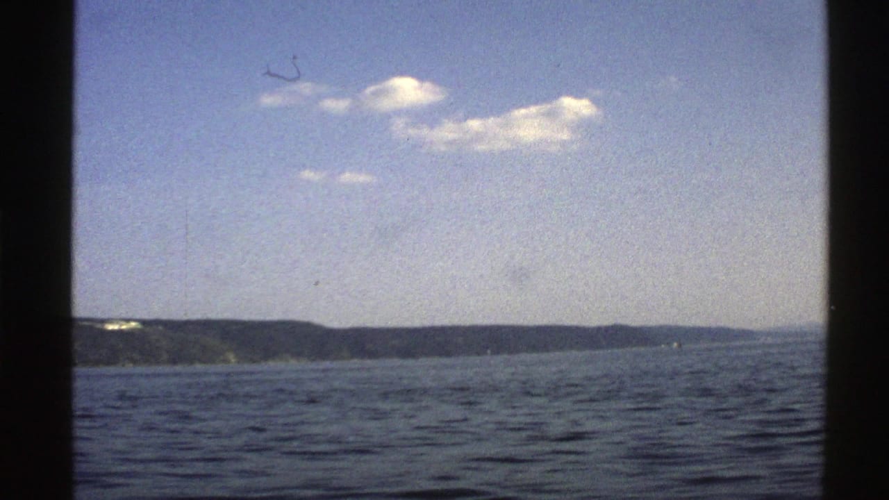 1976 Norway boat-view frame with rippled water, a long low shoreline, pale blue sky, and visible 8mm film edges.