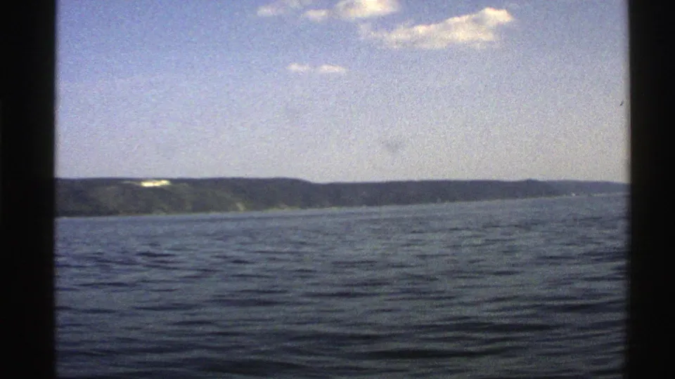1976 Norway boat-view still with calm dark water, a low coastal horizon, bright clouds, and black frame borders.