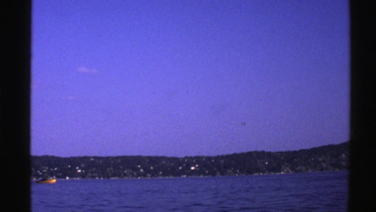 1976 Norway 8mm frame at dusk with blue water, a dark shoreline, scattered shore lights, and a small yellow boat.