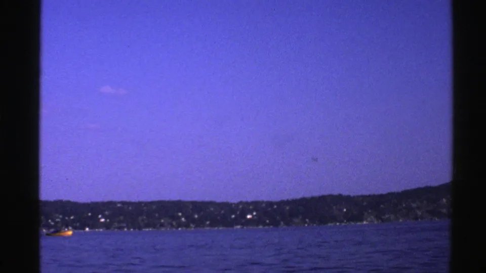 1976 Norway 8mm frame at dusk with blue water, a dark shoreline, scattered shore lights, and a small yellow boat.