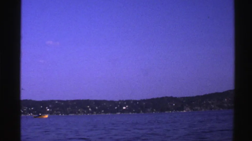 1976 Norway 8mm frame showing a blue dusk coastal view with shoreline lights, low hills, and a small yellow boat near frame edge.