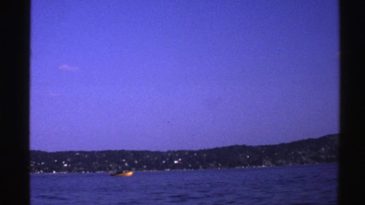 1976 Norway 8mm frame with a yellow boat traveling across blue water, seen against a distant wooded shoreline and open sky.