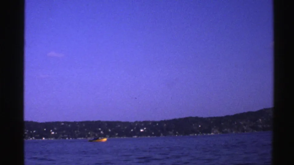 1976 Norway 8mm frame with a yellow boat traveling across blue water, seen against a distant wooded shoreline and open sky.