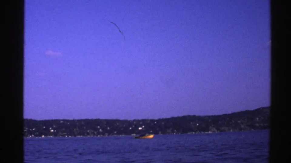 1976 Norway 8mm frame with the yellow boat near the far shoreline, surrounded by calm blue water and a broad clear sky.