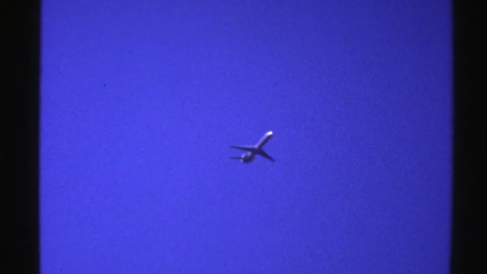 1976 Norway 8mm frame showing an airplane crossing a blue sky with strong film grain and color shift.