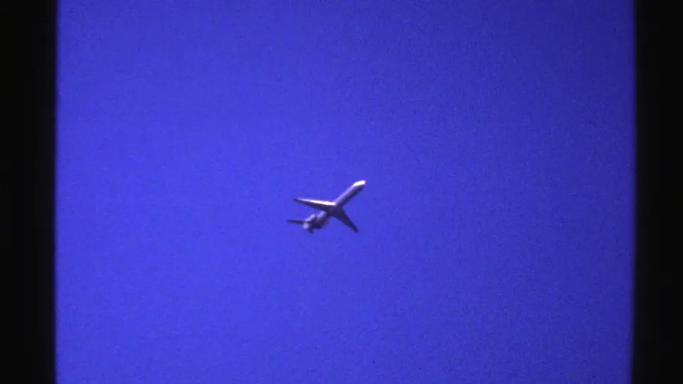1976 Norway 8mm frame showing a small passenger airplane crossing a cloudless blue sky, seen from below.