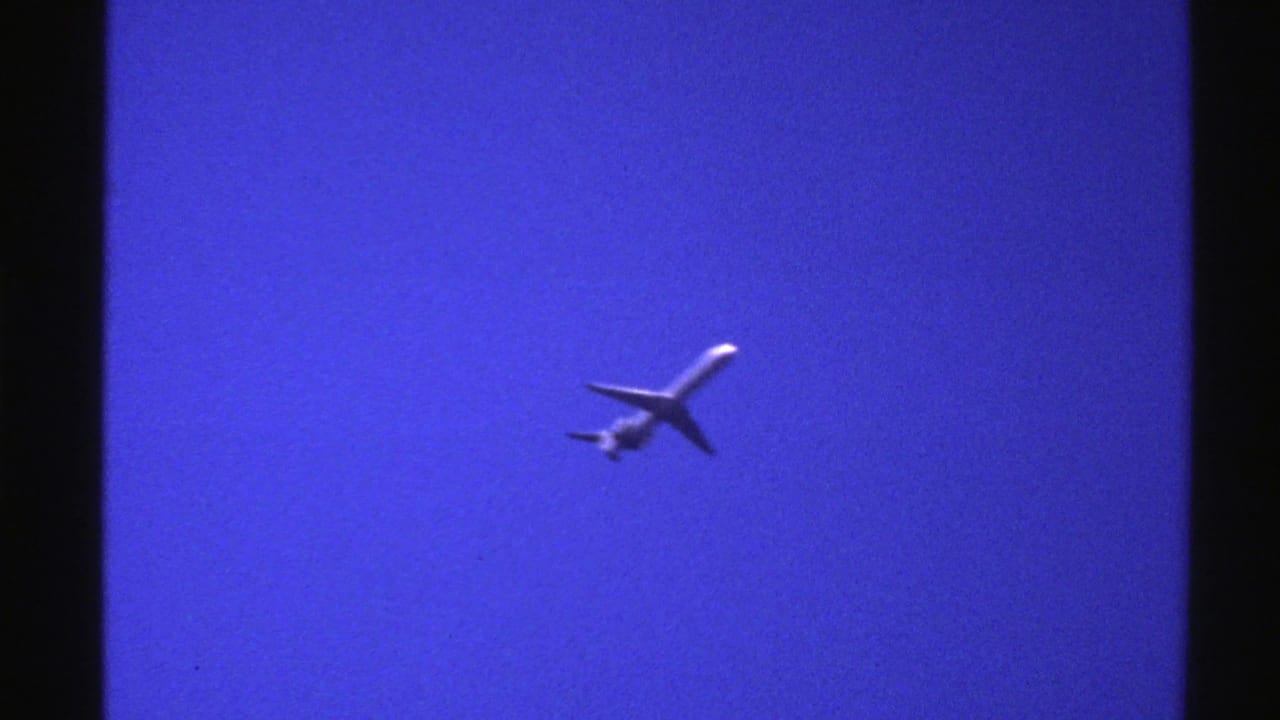1976 Norway home-movie frame with a distant passenger airplane angled across a saturated blue sky.