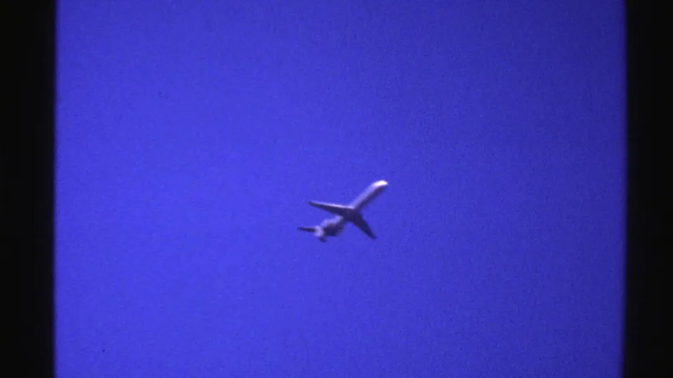 1976 Norway home-movie frame with a distant passenger airplane angled across a saturated blue sky.
