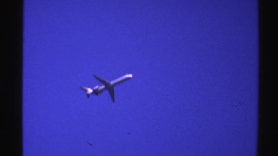 1976 Norway 8mm frame with a passenger airplane passing overhead, its underside and tail visible in the blue sky.