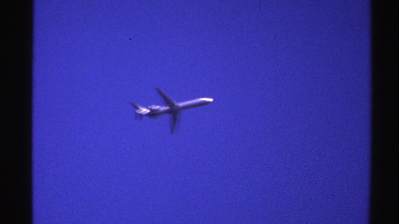A 1976 Norway 8mm frame shows a small commercial jetliner moving across a saturated blue sky with dark film borders visible.