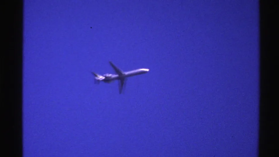 A 1976 Norway 8mm frame shows a small commercial jetliner moving across a saturated blue sky with dark film borders visible.