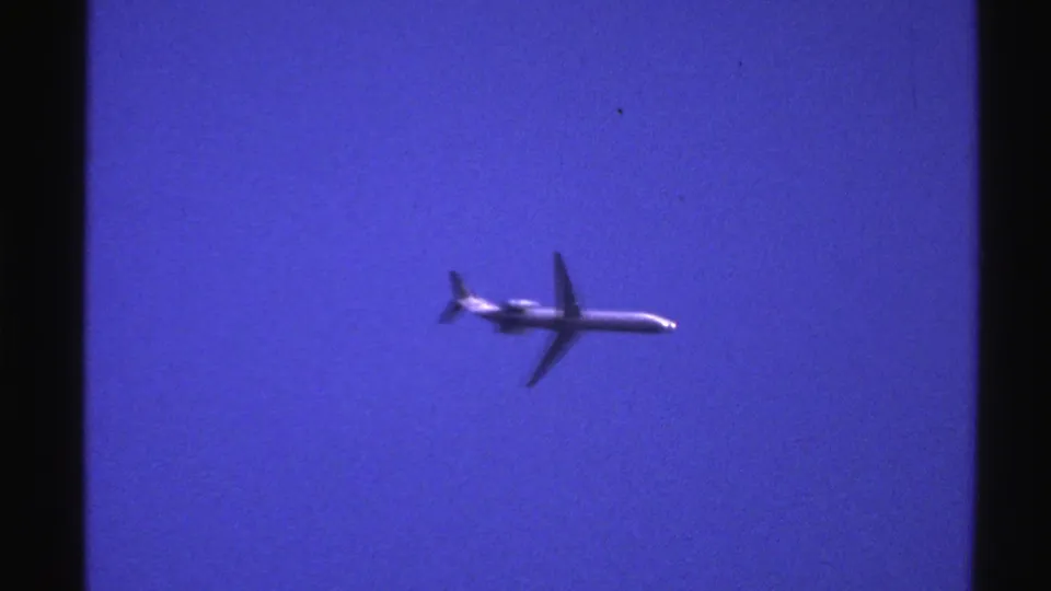 A white jet plane flies across a clear blue sky in Norway-sourced 1976 8mm home-movie footage.