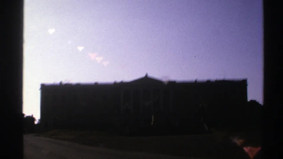 The camera holds on a silhouetted building facade with a roadway or drive in front, recorded on 1976 Norway-sourced 8mm film.