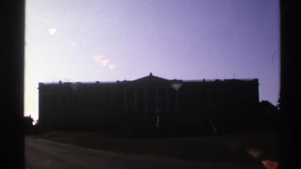 A dark classical-style building remains framed against a pale sky, with 8mm grain and flare visible in the shot.