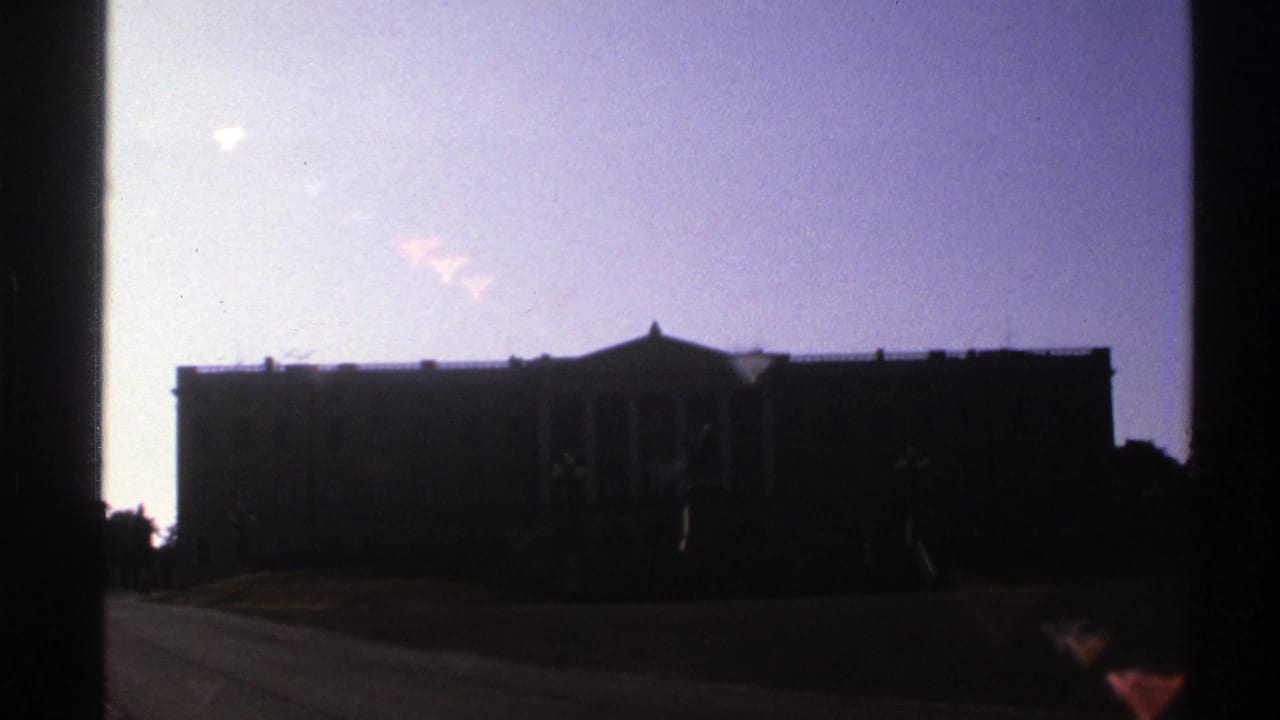 Dark silhouette of a columned building against a pale sky with lawn and path in front.