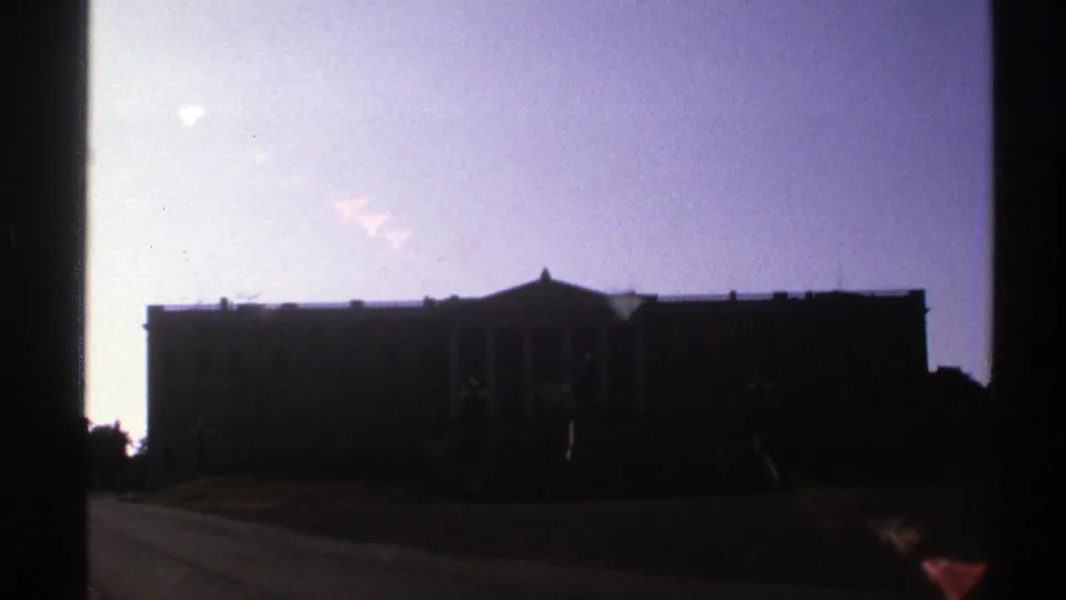 1976 Norway 8mm frame showing a silhouetted classical-style building with columns, lawn, paved path, and flare marks near the sky.