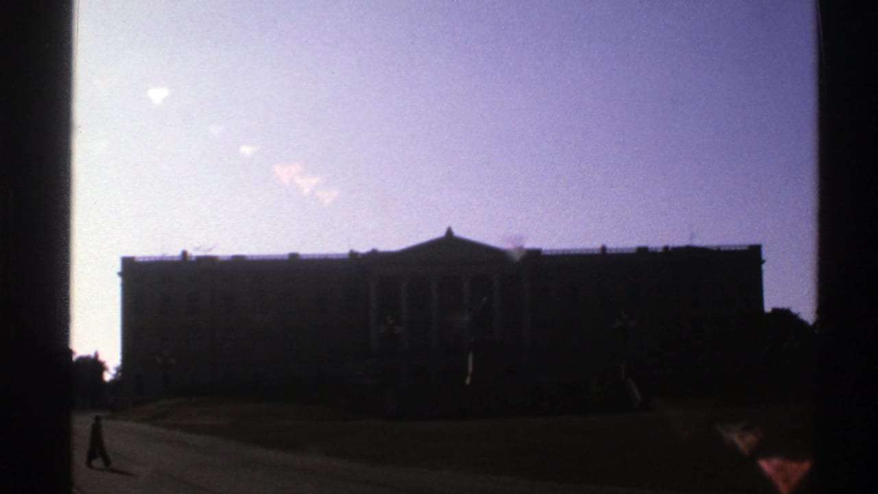 A distant person walks on a paved path before a dark columned building and pale sky.
