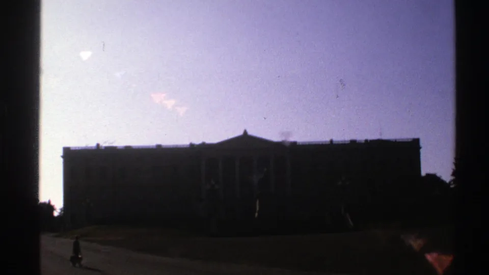 1976 Norway 8mm frame showing a distant walker near the edge of a paved path beneath a silhouetted columned building.