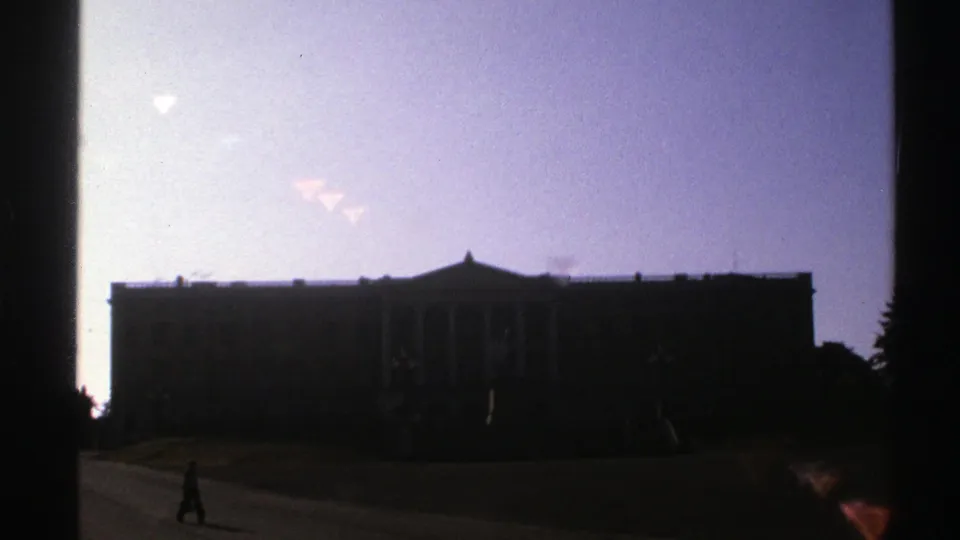 1976 Norway 8mm frame of a dark classical-style building, pale purple sky, lawn, and a small pedestrian on the path.