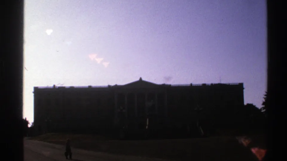 1976 Norway 8mm frame showing a dark formal building against a bright sky, with one distant pedestrian in front.