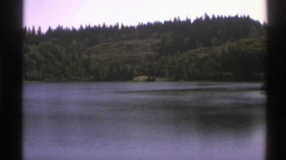 1976 Norway 8mm frame of lake water below a wooded hillside, with dark vertical film edges from the scan.