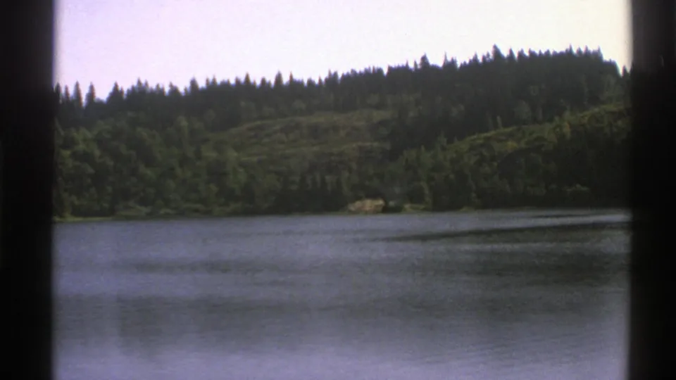 1976 Norway 8mm wide view across lake water toward dense trees and a low forested ridge.