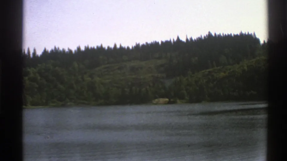 1976 Norway 8mm travel view of a calm lake with evergreen forest and wooded hills beyond, framed by dark film edges.