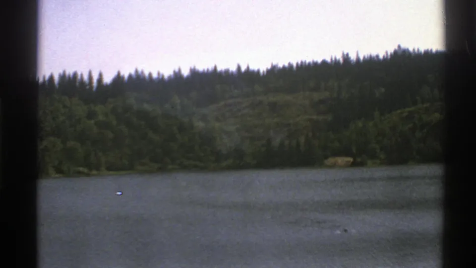 1976 Norway 8mm frame showing a lakeshore, dark evergreen forest, and rolling hills from a moving roadside viewpoint.