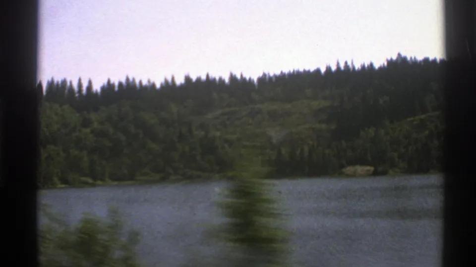 1976 Norway home-movie frame with motion-blurred roadside vegetation crossing a lake and forested hillside view.