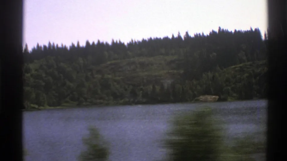 1976 Norway 8mm travel frame showing a lake, dense forest, low hills, and blurred roadside greenery from a moving vehicle.