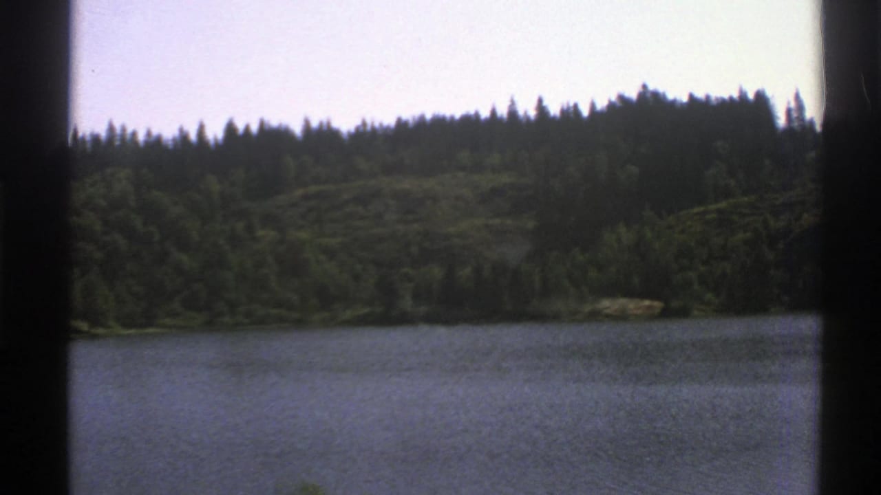 1976 Norway 8mm frame showing a calm lake or pond with wooded hills and evergreen shoreline.