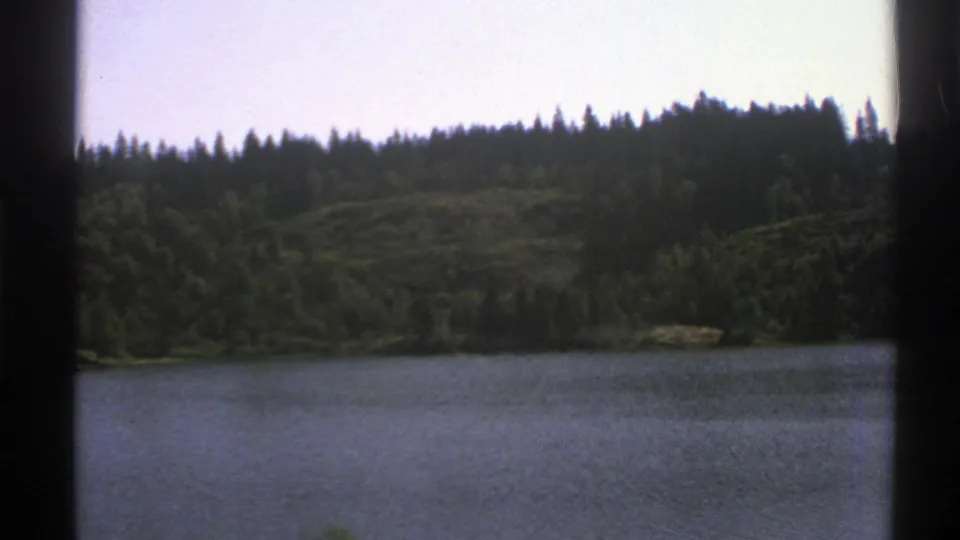 1976 Norway 8mm frame showing a calm lake or pond with wooded hills and evergreen shoreline.