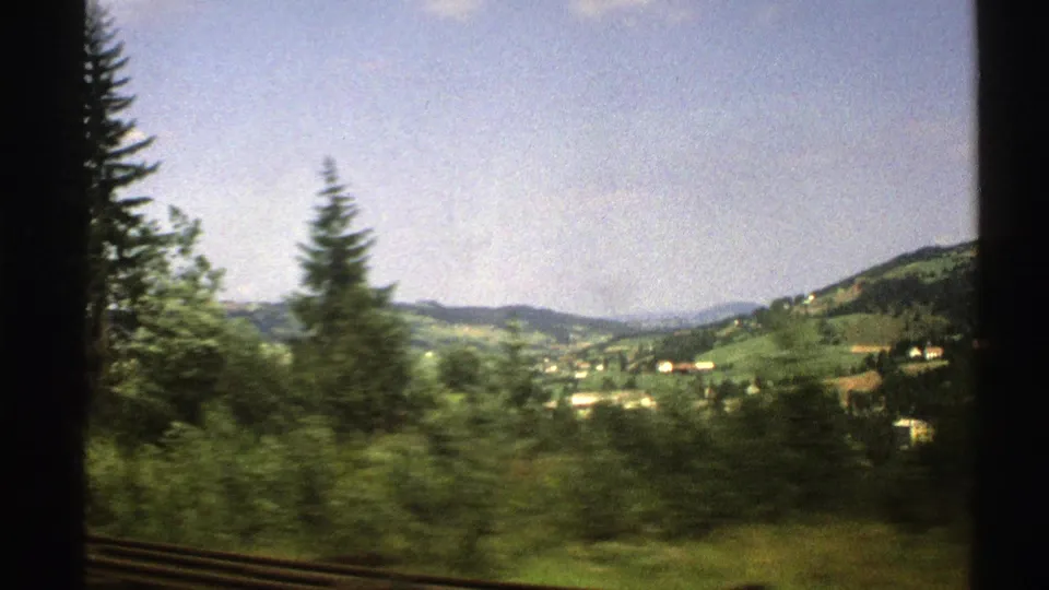 1976 Norway 8mm view of green hills, scattered houses, and conifer trees passing outside a train window.