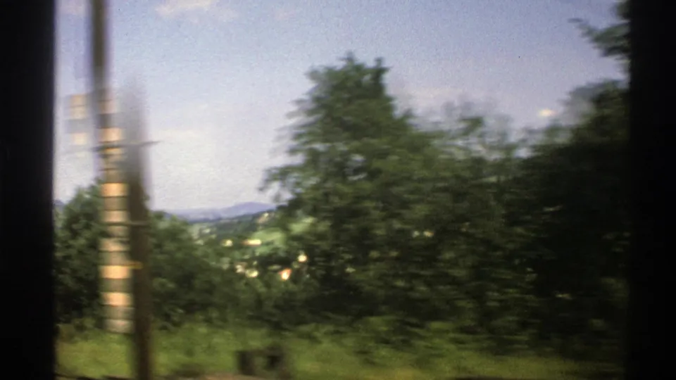 Moving train view in 1976 Norway with a passing pole, leafy trees, green valley slopes, and scattered buildings.