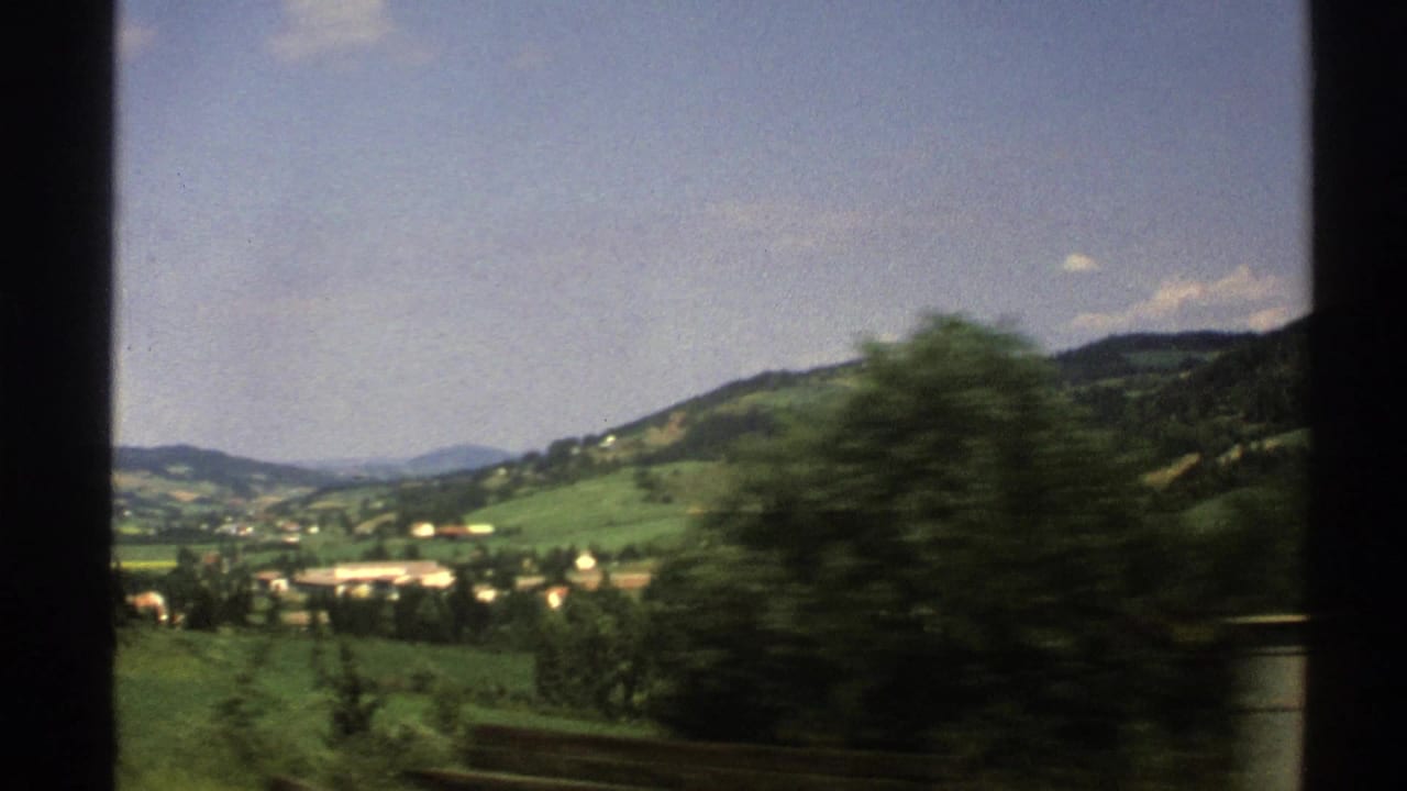 Train-window view of green hills, valley buildings, trees, and blue sky.