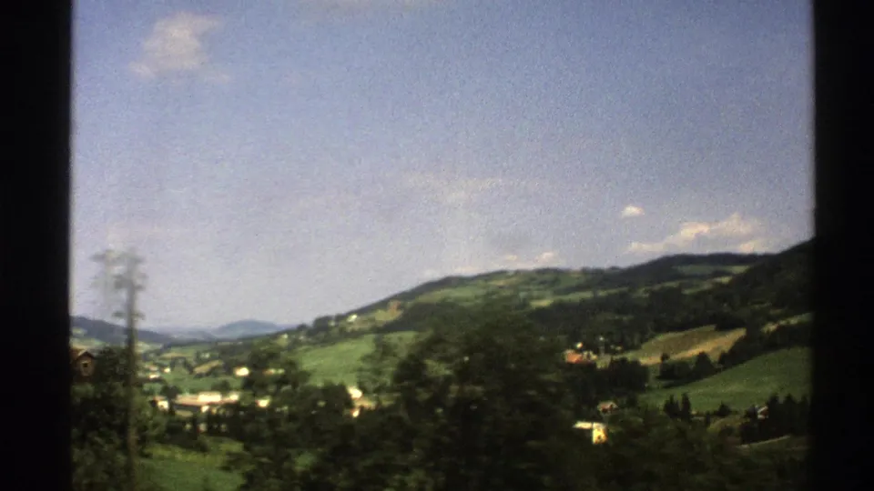 1976 8mm Norway view from a moving train, showing a utility pole, blurred trees, hillside buildings, and green slopes.