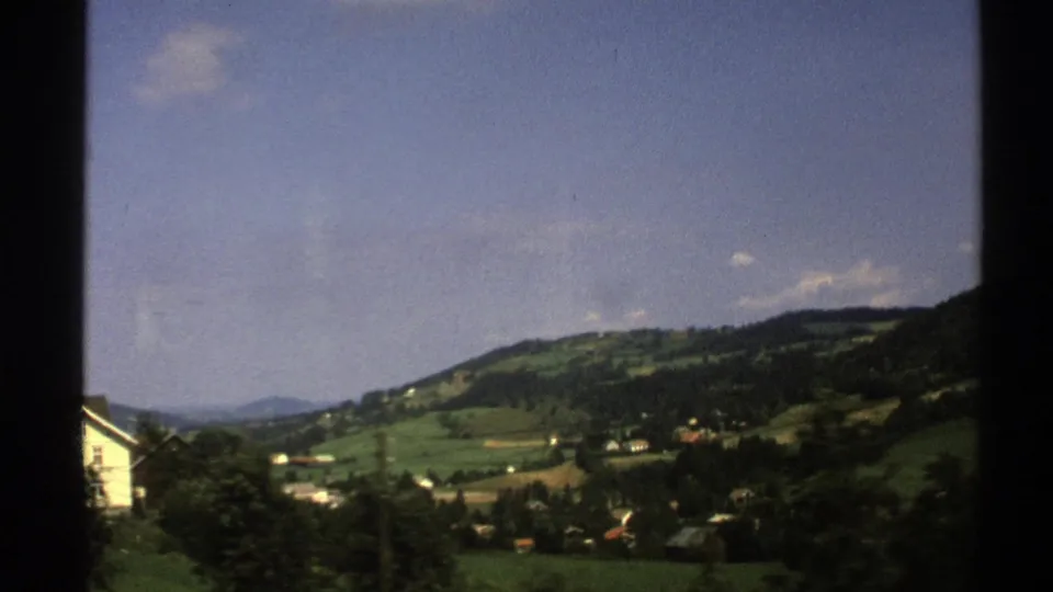 1976 Norway train-window frame with green valley fields, forested hillside, scattered buildings, and a bright blue sky.