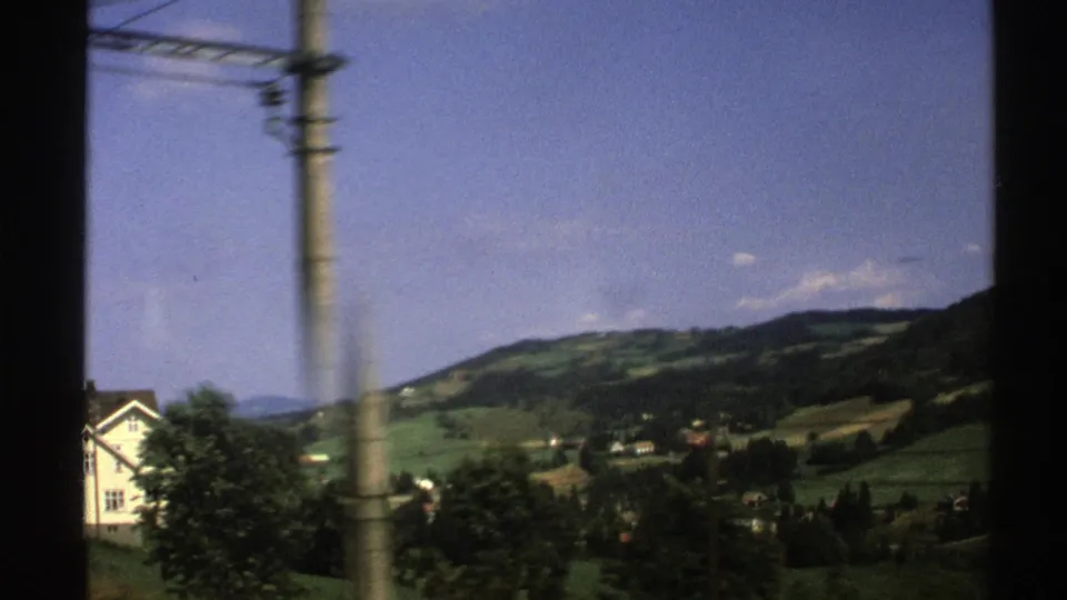 1976 Norway 8mm frame from a moving vehicle showing a white house, utility pole, wooded hills, green fields, and blue sky.