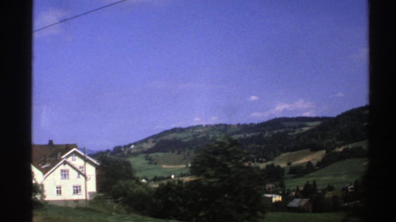 White house below green hills with trees, fields, overhead wire, and blue sky.