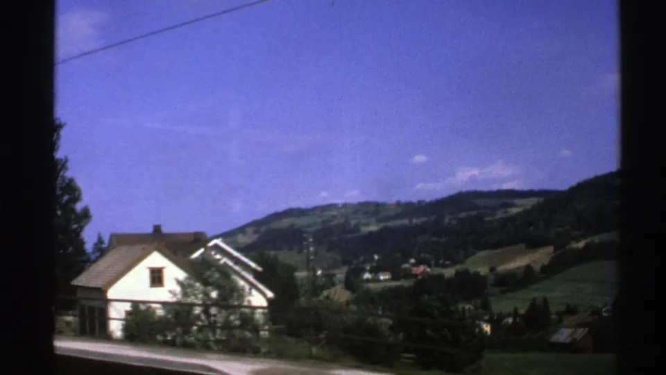 1976 Norway 8mm frame showing a white roadside house, fence, trees, open road edge, and layered green hills.